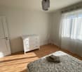 Bedroom with bed, white walls, wooden floor, white chest of drawers, white door, window with white curtains, a lamp hanging from the ceiling, and a chair