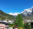 The image shows a scenic mountain landscape with snow-capped peaks in the background. There are several buildings and houses in the foreground, surrounded by lush greenery and trees. The sky is a clear, bright blue, creating a picturesque and serene setti