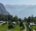 The image shows a scenic mountain landscape with a small village nestled in the valley below. The village consists of several traditional alpine-style houses surrounded by lush green forests and meadows. In the background, towering snow-capped peaks can b