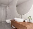 Modern bathroom with white ceramic tiles, round mirror, white vanity with wooden countertop, round basin, white toilet, and a glass shower stall with shower head and hand shower.