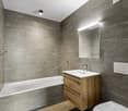 Bathroom with a bathtub, modern vanity, mirror, grey tiles, white porcelain sink, wooden cabinet, and a ceiling-mounted light