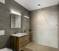 a modern and minimalistic bathroom with grey tiled walls, shower area with glass enclosure, wooden vanity with sink and mirror, a toilet