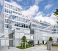 Multi-story office building with a modern, glass and concrete facade. The building has numerous windows and appears to be surrounded by greenery, including trees and landscaping. There is a parking area visible in the foreground of the image.