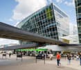 Large modern glass and steel commercial building with a covered bus station in the foreground. People are walking through the bus station area.
