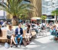 Outdoor seating area with palm trees, umbrellas, and people dining at tables. The area appears to be part of a commercial or office building complex with glass facades and a modern architectural style.