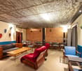 This image shows a cozy and eclectic living room space. The ceiling has a unique textured design with wooden panels. The walls are decorated with various artworks and sculptures. The room features a mix of comfortable seating options, including a red sofa