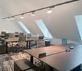 Modern office space with wooden desks, ergonomic chairs, track lighting, and large windows providing natural light. The room has a sleek and professional design aesthetic.