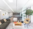 Spacious open-plan office with modern furniture, including a large sofa, armchairs, and a dining table. The space features large windows providing a view, and a potted plant adds a touch of greenery. The office appears to have a clean, minimalist design w