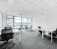 Spacious office with large windows, white walls, suspended ceiling with lighting fixtures, multiple desks and office chairs, carpeted floor