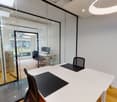Meeting room with glass walls, a large white desk, black chairs, a computer monitor on a desk, a large white ceiling light, and wooden flooring.