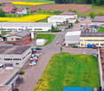 The image shows an aerial view of an industrial or commercial area. There are several buildings, including warehouses and office buildings, surrounded by paved parking lots and green spaces. The landscape includes fields with yellow flowers, forests, and