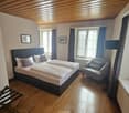 Spacious bedroom with wooden ceiling, large windows, gray armchair, and a king-size bed with a dark headboard. The room has a cozy and inviting atmosphere.