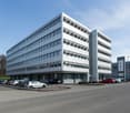 Multi-story office building with a modern, glass and steel facade. The building has several floors and a large parking lot in front of it.