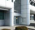 The image shows the exterior of a modern, multi-story building with a glass and metal facade. The building has a prominent address sign that reads 'Steinackerstrasse 29'. There are several large windows and glass panels on the facade, as well as some land