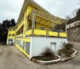 A two-story yellow building with a metal roof structure and balconies. The building has large windows and appears to be a commercial or community space. There is a small garden area in front of the building.