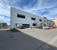 A large, modern commercial building with a flat roof and multiple windows. There is a parking area in front of the building with several cars parked. The building appears to be used for commercial or industrial purposes.