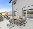 Modern two-story house with gray exterior, large windows, patio with outdoor furniture including a dining table and chairs, and a grassy yard.