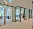 modern office space with glass walls, swivel doors, carpeted floor, desks, chairs, cabinets, and a trash bin