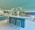 A white counter with a set of barstools and a blue backdrop with plants, along with tables and chairs. The space has a white floor and a green ceiling with hanging pendant lights.