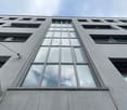 The image shows the exterior of a modern, multi-story building with a distinctive architectural design. The building features a large, vertical window panel that spans multiple floors, surrounded by a grid-like pattern of windows and concrete walls. The o