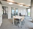 An empty, modern office with a gray carpeted floor, white walls, and ceiling lamps. It has a glass partition, white table and chairs, white cabinets, and two large windows. It also has two office desks with monitors.