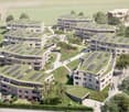 This image shows a large residential complex with multiple apartment buildings. The buildings have green roofs with solar panels installed, indicating the use of renewable energy. There are also visible balconies or terraces on the buildings, and parking