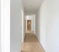 Long, narrow hallway with white walls and light hardwood flooring. The hallway leads to a doorway at the end, providing a view into another room.