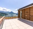 2-story wooden house with a large balcony overlooking a lake surrounded by mountains. The balcony has a tiled floor and metal railing. The house has a modern design with large windows and a scenic view of the lake and mountains.
