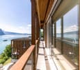 Multi-story wooden building with large windows and a balcony overlooking a lake and mountains in the background. The building has a wooden deck and railing system along the exterior.