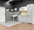 Empty room with a white door, dark gray walls, light gray floor, white sink with a faucet, two white cabinets, a wall-mounted tissue dispenser, and a mirror