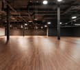 Spacious room with wooden floors and black walls, several pillars, ceiling lamps and ceiling pipes.
