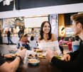 The image shows a group of people dining at a restaurant. A smiling young woman is seated at a table, holding a burger and surrounded by various food items. Other patrons can be seen in the background, creating a lively atmosphere.