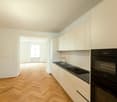 An empty kitchen with white cabinets, wooden flooring, a double sink, and black countertops.