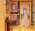 Ornate and decorative interior with wooden walls, checkered floor tiles, antique furniture, and framed artwork on the walls. The room appears to lead into another room or hallway through an ornate doorway.