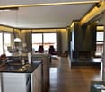 The image shows an open-plan living space with a kitchen area, a living room with a fireplace, and large windows providing a view of the outdoors. The space has a modern, rustic design with wooden floors, stone walls, and warm lighting.
