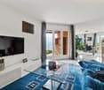 Spacious living room with large windows, glass coffee table, blue sofa, TV, and decorative wall art. The room has access to a balcony or terrace and features a fireplace.