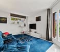 Spacious living room with large windows, blue velvet sofa, glass coffee table, wall-mounted TV, shelves, and access to a balcony or terrace.