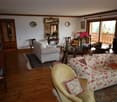Spacious living room with hardwood floors, white walls, and wooden beams. The room features a floral patterned sofa, a round wooden coffee table, and a large mirror on the wall. There is also a balcony visible through the window, providing natural light a