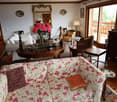 Cozy living room with wooden furniture, floral patterned sofa, ornate mirror, and a vase of pink flowers on a round wooden table. The room has large windows overlooking a balcony or terrace.
