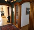 A hallway with a decorative rug, wooden door frames, and a vintage chandelier. A toilet and a shelf with decorative items are visible through an arch doorway. An open window and a TV are in the back.