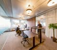 large open space with desks, office chairs, standing desks, people working on laptops, white curtains, carpeted floor, windows with view to outside