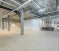 Large open office space with concrete ceiling, exposed pipes and ductwork, concrete floor tiles, and glass-enclosed meeting rooms or offices. The space appears to be a modern, industrial-style office environment.