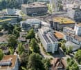 Urban aerial view of residential and commercial buildings, green rooftops, solar panels on some buildings, greenery, roads, and vehicles.