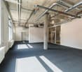 Large open office space with concrete ceiling, exposed pipes and ductwork, gray carpeted floor, white walls, and a door leading to another area.