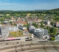 Aerial view of a city with a mix of modern and historic buildings, surrounded by lush green hills and forests. The city has a train station and parking facilities visible in the foreground.