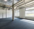 Large open office space with exposed ceiling, concrete columns, and large windows overlooking the exterior. The space has a gray carpet floor and appears to be in a commercial or industrial building.