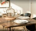 Dining room with wooden table, 6 chairs, plates, wine glasses, cutlery, and a glass vase