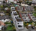 Modern houses in hilly area, with raised ground floor, several balconies, terraces, solar panels on roofs, parking places, surrounded by greenery