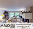 white living room with fireplace, dining area, white sofa, armchairs, desk, coffee table, chandelier, paintings, lamp, dining table, chairs, kitchen, picture frame, television, books, and basket