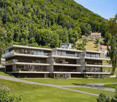 Modern multi-story building with glass walls, balconies, and solar panels on the roof, set in a lush green landscape with a mountain in the background.